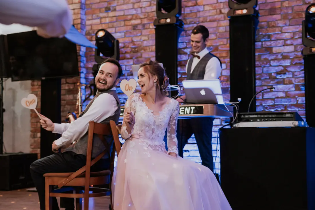 Para na weselu podczas zabawy z tabliczkami, w tle DJ przy konsolecie
